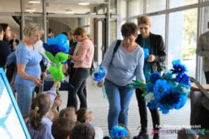 W holu Chatki Żaka kobiety rozdają dzieciom kwiaty wykonane z bibuły w różnych odcieniach niebieskiego koloru