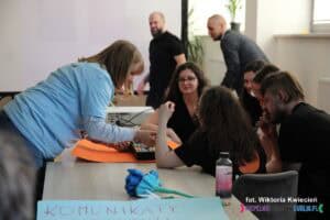 Pani Katarzyna Siedlecka - psycholog i pedagog specjalny, koordynator wolontariatu rówieśniczego Fundacji ALPHA, pochyla się nad stołem, przy którym siedzi grupa młodych osób