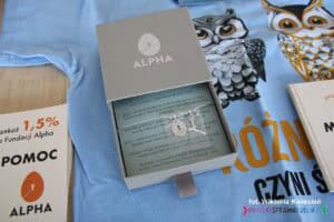 W szarym pudełeczku leży srebrna zawieszka na łańcuszku w kształcie jajka i klucza, w tle na niebieskim papierze opisano symbolikę biżuterii