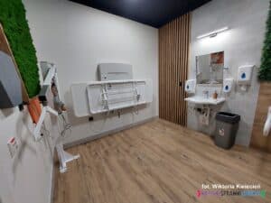 Widok na zamontowane na różnych ścianach: składany przewijak, podnośnik oraz umywalkę