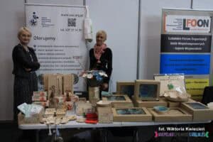 Strefa NGO na Targach Pracy: stoisko ZAZ przy Stowarzyszeniu 'Promyk' w Łukowicy. Do zdjęcia pozują dwie reprezentantki placówki. Na stole przed nimi ułożono ceramikę opakowaną w kartonowe pudełka-zestawy prezentowe oraz wyroby z grawerowanego drewna