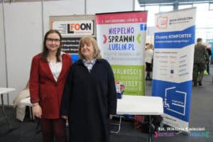 Targi Pracy: dwie kobiety pozują przy stoisku Lubelskiego Forum Organizacji Osób Niepełnosprawnych - Sejmik Wojewódzki., od lewej: Pani Wiola Story, Psycholog CIPON oraz Pani Alicja Jankiewicz, Prezes Honorowa LFOON; w tle banery LFOON-u Biuletynu oraz Koalicji Przewijamy Polskę