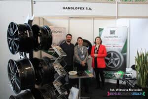 Targi pracy; stoisko firmy Handlopex S.A. Od lewej stoi mężczyzna, obok niego dwie kobiety — wszyscy zwróceni w stronę aparatu. Po lewej stronie kadru widoczny jest stojak prezentujący czarne felgi, a obok niego drugi stojak z katalogami firmowymi