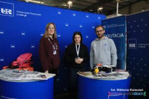 Stoisko Uniwersytetu Rzeszowskiego. na targach pracy. Trzy osoby stoją obok siebie: dwie kobiety i mężczyzna. Po obu stronach znajdują się stoliki z materiałami promocyjnymi