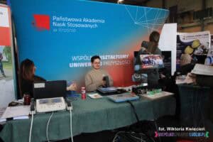 Targi pracy: na stoisku Państwowej Akademii Nauk Stosowanych w Krośnie siedzą dwie osoby: kobieta i mężczyzna. Przed nimi na stole leżą modele robotów lub urządzenia związane z robotyką, stanowiące część prezentacji