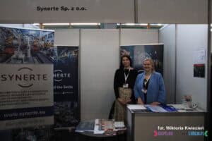 Na stoisku firmy Synerte Sp. z o.o. dwie kobiety uśmiechają się do aparatu, pozując do zdjęcia. Na stoliku przed nimi znajdują się materiały promocyjne, takie jak broszury i ulotki