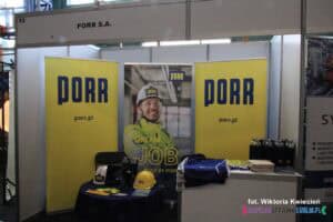 Stoisko firmy budowlanej Porr S.A. na targach pracy. Na stołach ułożone materiały promocyjne, w tym żółty i biały kask budowlany