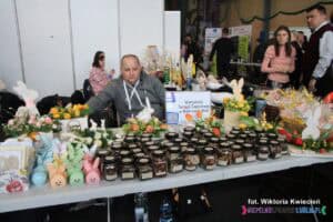 Na stoisku Warsztatów Terapii Zajęciowej w Bobrowej Woli podczas targów pracy, siedzi mężczyzna. Na stole przed nim widoczna jest różnorodność rękodzieła takich jak ozdoby wielkanocne i szklane słoiczki wypełnione ziołami