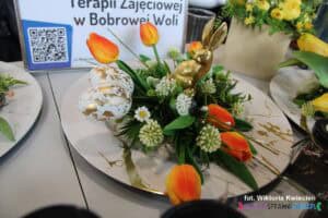 Stroik wielkanocny na talerzu ze złotym zającem i pomarańczowymi tulipanami na stoisku Warsztatów Terapii Zajęciowej w Bobrowej Woli