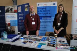 Stoisko Głównego Punktu Informacyjnego Funduszy Europejskich w Rzeszowie na Targach Pracy. Na stoisku mężczyzna i kobieta pozują do zdjęcia, a przed nimi na stoliku leżą ulotki i materiały promocyjne