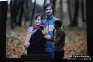 Podczas prelekcji Pana Jana Meli w tle na telebimie wyświetlone jest zdjęcie, na którym prelegent stoi w jesiennym lesie z młodą kobietą i niemowlęciem. Prelegent trzyma mikrofon w lewej ręce i patrzy za siebie, na ekran wyświetlający tę fotografię, skupiając uwagę publiczności na wspomnianym obrazie