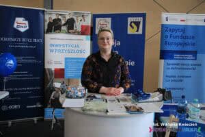 Na stoisku Podkarpackiej Wojewódzkiej Komendy Ochotniczych Hufców Pracy w Rzeszowie widoczna jest kobieta pozująca do zdjęcia. Przed nią na stoliku ułożone są liczne materiały promocyjne, takie jak broszury, ulotki i plakaty, które informują o działalności Hufców Pracy