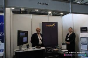 Stoisko firmy Grinea na Targach Pracy. Dwie kobiety pozują do zdjęcia. Na stole po lewej stoi telewizor z włączoną ofertą firmy a po prawej stojak z ulotkami
