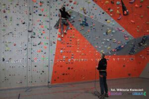 Na ściance wspinaczkowej kobieta wspina się w górę, a u dołu mężczyzna ją asekuruję, trzymając linę