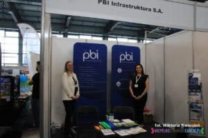 Stoisko PBI Infrastruktura S.A. na Targach Pracy. Dwie kobiety pozują, a na niskim stoliku przed nimi ułożono materiały informacyjne i promocyjne.