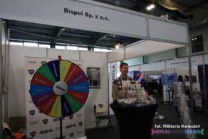 Na stoisku firmy BISPOL Sp. z o.o. podczas Targów Pracy mężczyzna stoi przy stole, na którym zaprezentowane są produkty firmowe. Po lewej stronie stoisko wyposażono w grę przypominającą Koło Fortuny, która oferowała zabawę i możliwość wygrania nagród