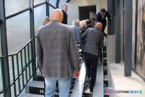 Mężczyźni asystujący osobie poruszającej się na wózku przy mobilnych rampach teleskopowych, umieszczonych na schodach prowadzących do Centrum Historii Sportu