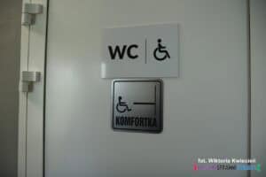 Drzwi wejściowe z plakietkami informującymi o toalecie dla osób z niepełnosprawnością i oznaczeniem Komfortka
