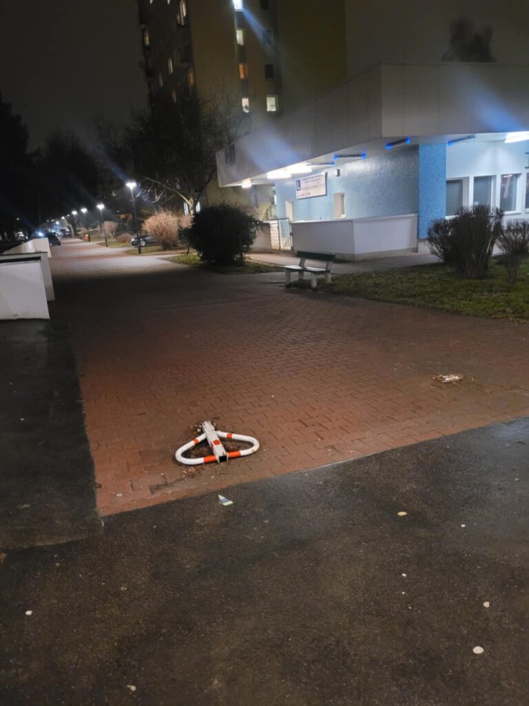 Wieczór. Fragment chodnika stanowiący podjazd pod Dom Kultury ze złożoną blokadą wjazdu. W tle fragment oświetlonego pasażu handlowego, trawnik, krzewy oraz blok mieszkalny