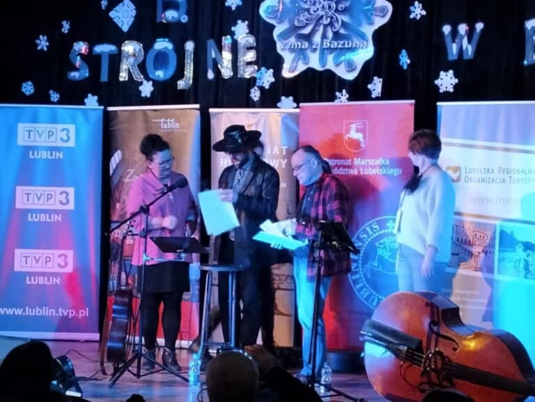 Moment wręczenia wyróżnienia w „Konkursie jednego wiersza”. Na scenie stoi zdobywczyni nagrody, Dorota Rudzik, ubrana w różowy żakiet i ciemną spódnicę, obok niej członkowie jury, m.in. organizator festiwalu Józef Bilski. Z tyłu – banery sponsorów oraz fragment dekoracji sceny, część napisu „Strojne w biel” i śnieżynki z kartonu. Przed osobami stoi sprzęt nagłaśniający oraz instrumenty – gitara i kontrabas