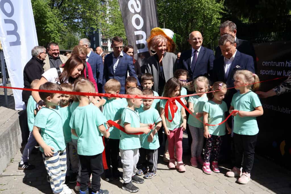 Grupa dzieci i dorosłych stoi przed czerwoną wstęgą zawiązaną na kokardę, tuż przed otwarciem Centrum Historii Sportu. W tle widać zieleń drzew. Uczestnicy uśmiechają się.