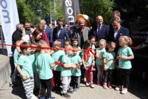 Grupa dzieci i dorosłych stoi przed czerwoną wstęgą zawiązaną na kokardę, tuż przed otwarciem Centrum Historii Sportu. W tle widać zieleń drzew. Uczestnicy uśmiechają się.