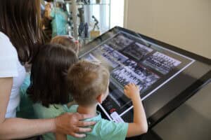 Dzieci pod opieką dorosłej kobiety oglądają zdjęcia wyświetlone na poziomym ekranie multimedialnym