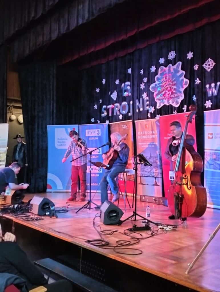 Na scenie trzej artyści festiwalu: z lewej skrzypek Maciej Błyszczak (bordowe spodnie, kraciasta koszula), w środku gitarzysta Jan Błyszczak „Mufka” (niebieskie jeansy i bluza), oraz kontrabasista (ciemny sweter i koszulka). Przed nimi sprzęt nagłaśniający, w tle banery sponsorów. Zza kulis wygląda organizator Józef Bilski (czarne ubranie, skórzana kurtka, kowbojski kapelusz).