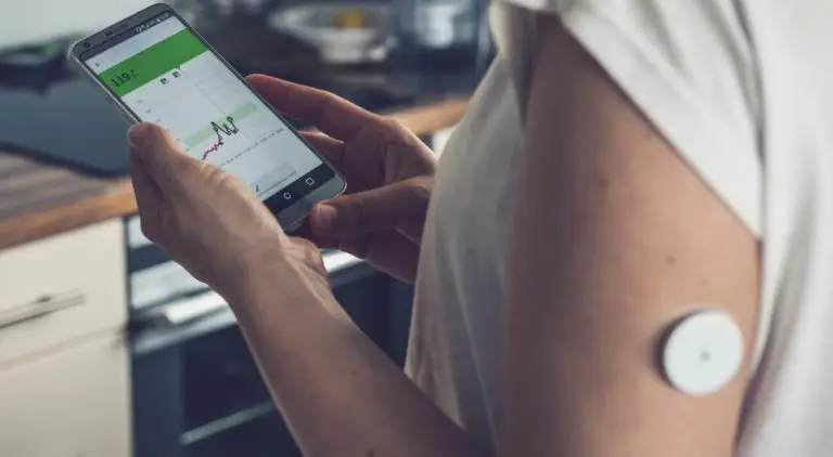 Kobieta trzymająca smartfon, na którym wyświetlany jest wykres, ma na górnym ramieniu przymocowany biały, okrągły czujnik monitorujący dane glukozy.