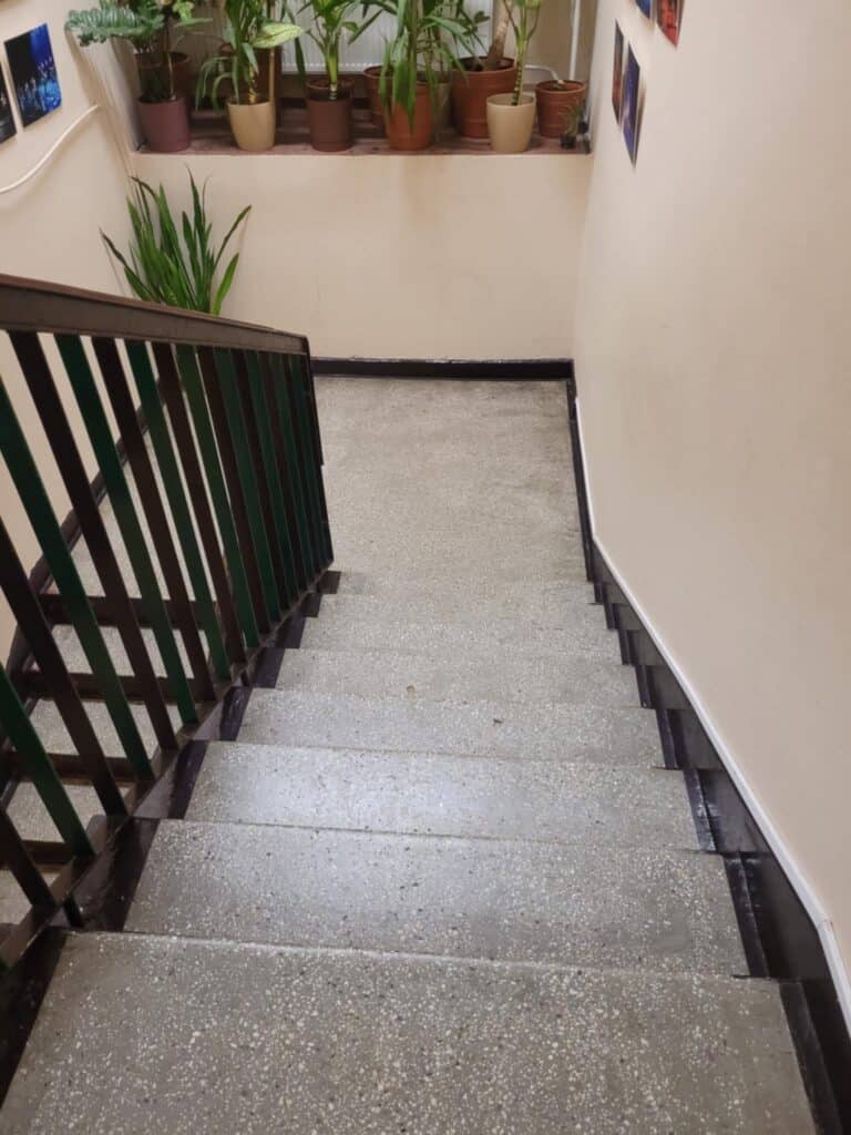 Schody prowadzące do toalety oraz szatni w Klubie Osiedlowym, kondygnacja biegnąca w dół. Schody zrobione są z lastryko, mają szczebelkową poręcz w kolorze brązowym. Na podeście schodów stoi kwiatek doniczkowy, widać fragment okna i parapetu z roślinami doniczkowymi w brązowawych doniczkach. Po obu stronach – fragmenty zdjęć na ścianach. Ściany i schody w kolorze beżowym.