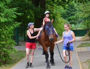 Dziecko w kasku jedzie na brązowym koniu po ścieżce, obok idą dwie kobiety – jedna prowadzi konia, a druga stabilizuje dziecko. W tle widoczne są drzewa
