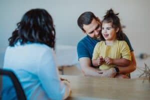 Mężczyzna trzyma na kolanach uśmiechniętą dziewczynkę z Zespołem Downa, siedząc przy stole. Naprzeciwko nich siedzi kobieta tyłem do aparatu, która angażuje się w rozmowę.