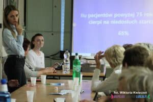 Sala konferencyjna podczas prelekcji. Przy kilku stołach siedzą uczestnicy spotkania, zwróceni twarzami w stronę prelegentki, która jako jedyna stoi. Na stołach znajdują się jednorazowe kubeczki i butelki z wodą. W tle, na dużym ekranie, widoczny jest slajd z pytaniem: 'Ilu pacjentów powyżej 75 roku życia cierpi na niedosłuch starczy?' oraz odpowiedzią: '65%'