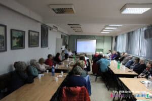 Widok na salę konferencyjną w trakcie trwającego spotkania. Uczestnicy siedzą przy długich stołach, skierowani w stronę końca sali, gdzie znajduje się monitor multimedialny z wyświetlaną prezentacją