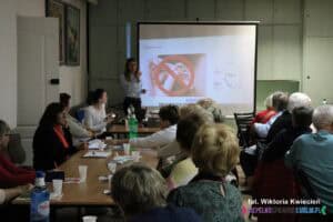 Uczestnicy spotkania siedzą przy długich stołach, zwróceni w stronę prelegentki stojącej obok ekranu multimedialnego. Na ekranie wyświetlony jest slajd dotyczący higieny uszu.