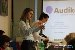 Prelegentka Wioleta Tatara gestykuluje podczas wypowiedzi. W tle widać pochyloną nad stołem ekspertkę Martę Pomianowską-Momont oraz slajd z danymi teleadresowymi gabinetu Audika