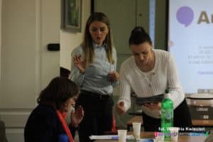 Prelegentki stoją przy stole, obok siedzącej uczestniczki. Jedna gestykuluje, a druga pochyla się, trzymając stos voucherów i wyciągając rękę w stronę uczestniczki