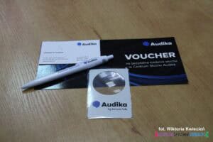 Na blacie w kolorze drewna leżą gadżety reklamowe z logo i nazwą Audika: voucher, długopis oraz lupka powiększająca