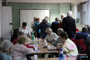 Widok na salę konferencyjną: część uczestników siedzi przy stołach, a część ustawiła się w około dziesięcioosobowej kolejce do badania wnętrza ucha przy pomocy otoskopu