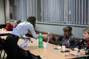 Prelegentka trzyma w lewej ręce naręcze smyczy reklamowych, a prawą podaje gadżet jednej z uczestniczek siedzącej przy stole