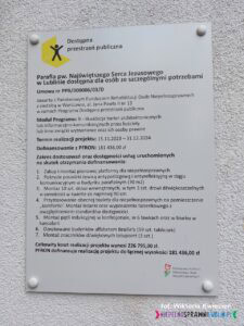 Na białej elewacji budynku przymocowano tablicę informacyjną, na której znajdują się szczegóły dotyczące zrealizowanego projektu „Dostępna przestrzeń publiczna”