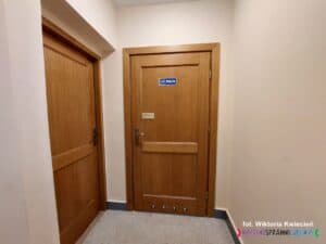 Drewniane, zamknięte drzwi. W górnej części drzwi znajduje się granatowo-biała tabliczka z napisem „Toaleta”. Niedaleko klamki znajduje się złota tabliczka z czarnym napisem „Toaleta – niepełnosprawni” oraz oznaczenie w alfabecie Braille’a