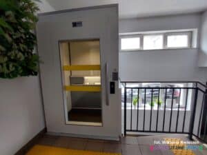 Szklane drzwi wejściowe do windy znajdujące się na pierwszym piętrze, oznaczone żółtymi kontrastowymi pasami poziomymi