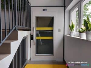 Szklane drzwi wejściowe do windy znajdujące się na parterze, oznaczone żółtymi kontrastowymi pasami poziomymi