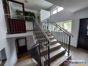 Siedem schodków łączących dwa poziomy w budynku parafialnym. W tle widoczny jest dźwig osobowy, którym można poruszać się między poziomami