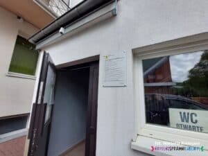 Niski budynek z białą elewacją. Widoczne są otwarte drzwi, a w oknie znajduje się plansza z napisem „WC otwarte”. Na ścianie pomiędzy drzwiami a oknem wisi tablica informacyjna, na której znajdują się szczegóły dotyczące zrealizowanego projektu „Dostępna przestrzeń publiczna”.
