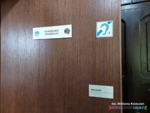 Ciemnobrązowe drzwi prowadzące do kancelarii parafialnej, oznaczone tabliczkami informacyjnymi, w tym symbolem ucha z literą „T”, wskazującym na obecność pętli indukcyjnej dla osób niedosłyszących