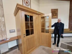 Wnętrze kościoła. Drewniany, zamykany konfesjonał z dwoma osobnymi wejściami — po lewej dla spowiednika, po prawej dla penitenta. Przy otwartych drzwiach po stronie penitenta stoi proboszcz, ks. Waldemar Sądecki. Na przeszklonych drzwiach widoczne są dwa oznaczenia: u góry granatowy symbol pętli indukcyjnej (ucho z przekątną linią i literą „T”), a nad klamką tabliczka z czarnodrukiem i alfabetem Braille’a