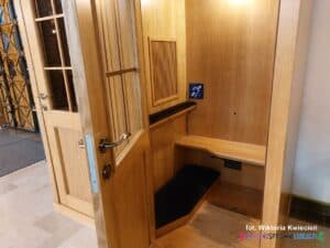 Wnętrze zamykanego konfesjonału po stronie penitenta. Na ścianie widoczny granatowy symbol pętli indukcyjnej — ucho z przekątną linią i literą „T”