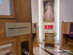 Kaplica spowiedzi. Ostrość aparatu skupiona na drzwiach z tabliczką w czarnodruku i alfabecie Braille’a. W tle, w mniejszej ostrości, widać wnętrze kaplicy z konfesjonałami i klęcznikiem stojącym po środku. Na ścianie wisi obraz Jezusa Miłosiernego a pod nim umieszczono relikwie dwóch polskich błogosławionych spowiedników: ks. Michała Sopoćki i o. Honorata Koźmińskiego